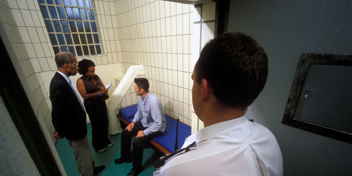 Two independent custody visitors in a police cell talked to a detained person, while a custody officer stands outside the open door