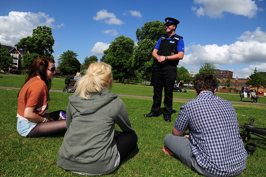 a group of people sat on grass, talking to a police officer standing in front of them