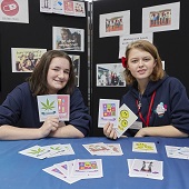 Two Youth Commission members hold postcards, with more on a bue tablecloth in front of them; pictures are displayed on a black board behind them.