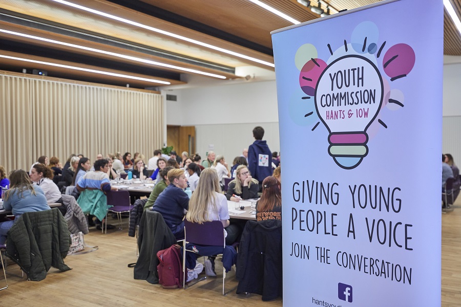 Lots of people seated in a hall, with a banner for the Youth Commission in the foreground which has their logo and says "giving young people a voice" and "join the conversation"