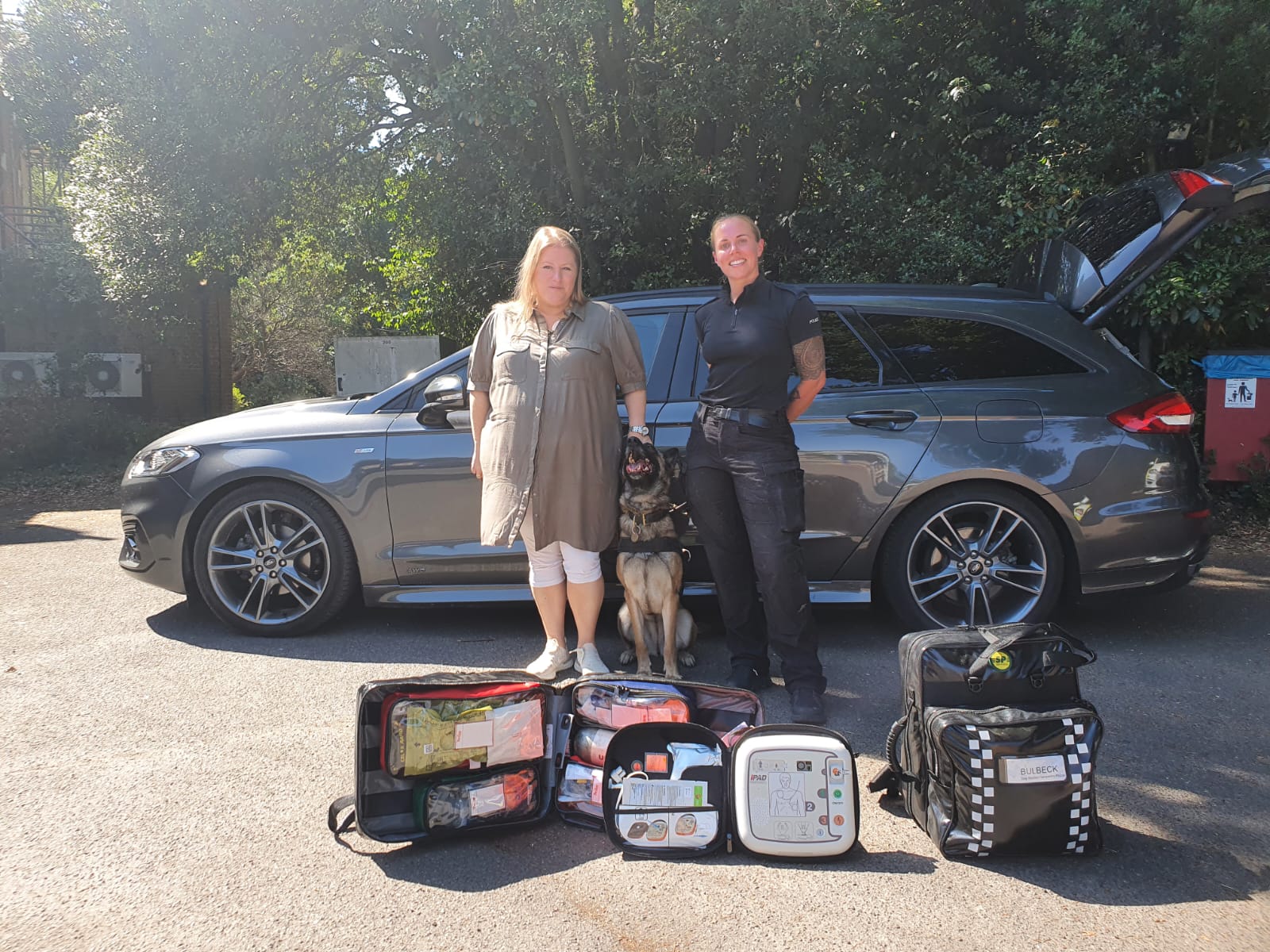 Donna Jones standing next to a police dog and police officer behind several bag of first aid kit, in front of a car. Looking happy