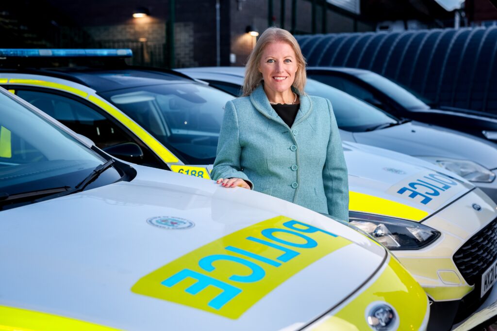 PCC smiling in a blue jacket with her hand on the bonnet of a police car.