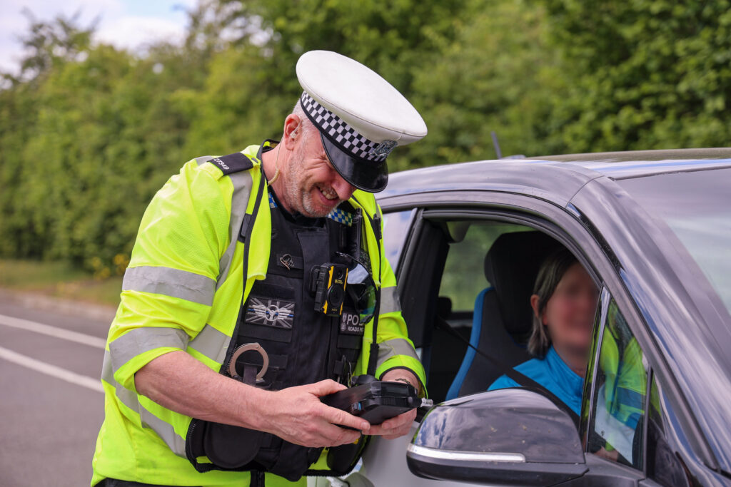 A uniformed traffic police officer looking at the reading on a portable Nitrous Oxide breathalyser. He is stood next to a car, open window next to the driver.