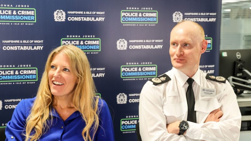 PCC Donna Jones and Chief Constable Alexis Boon, both smiling, sat in front of a board with repeated OPCC and Constabulary logos on it.
