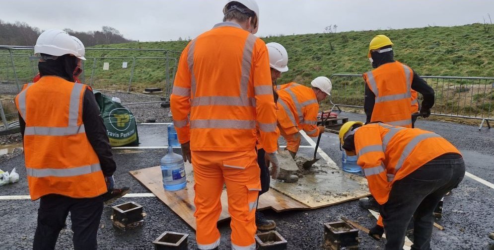 A group of six people, all in high visibility jackets and hard hats, working together with building materials, on a tarmac area.
