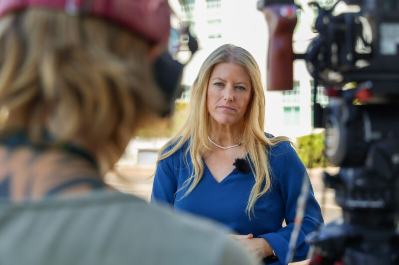 PCC Donna Jones in a blue dress with a serious expression on her face as she gives an interview. There is an out of focus camera operator and tv camera on a tripod in the foreground.