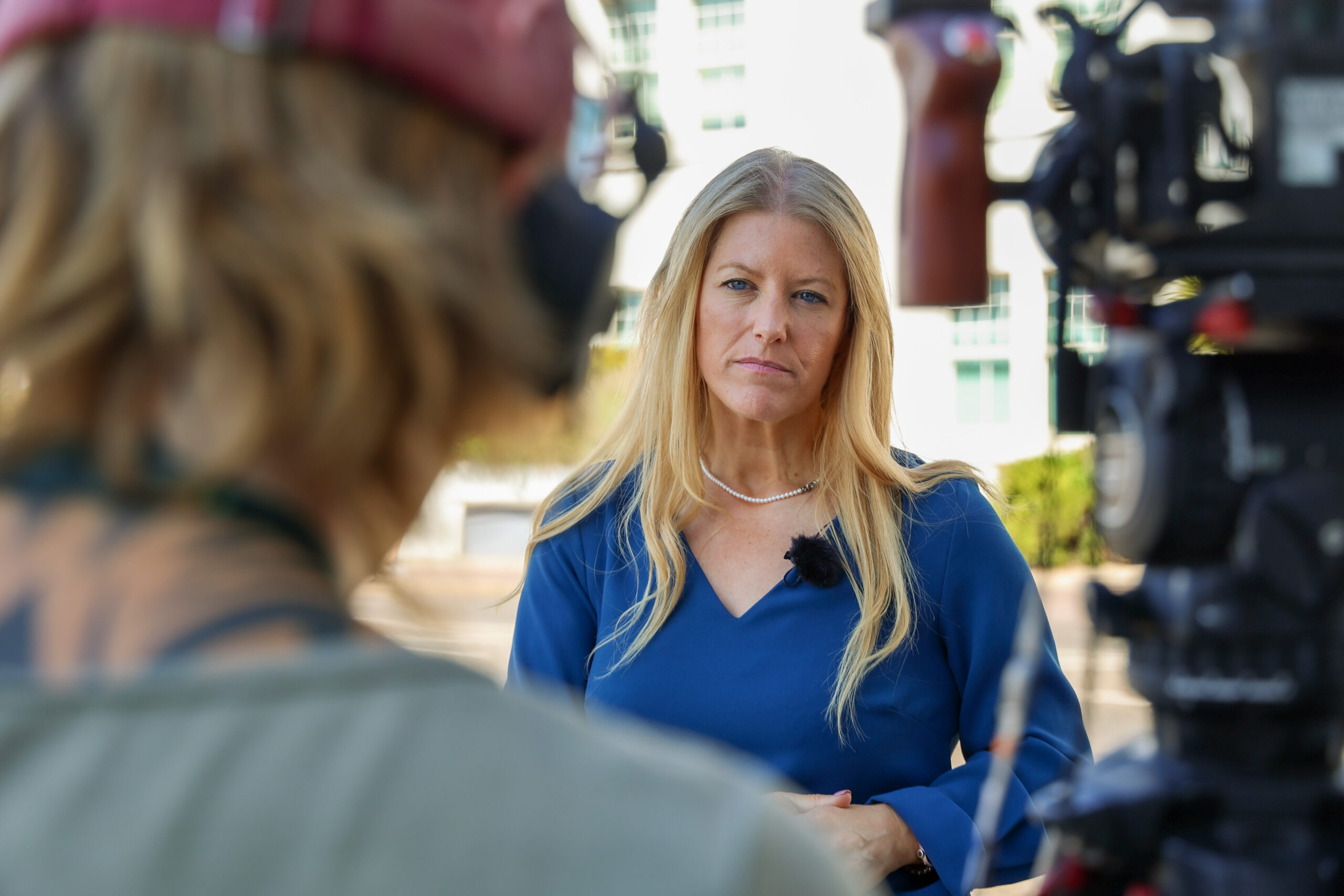 PCC Donna Jones in a blue dress with a serious expression on her face as she gives an interview. There is an out of focus camera operator and tv camera on a tripod in the foreground.