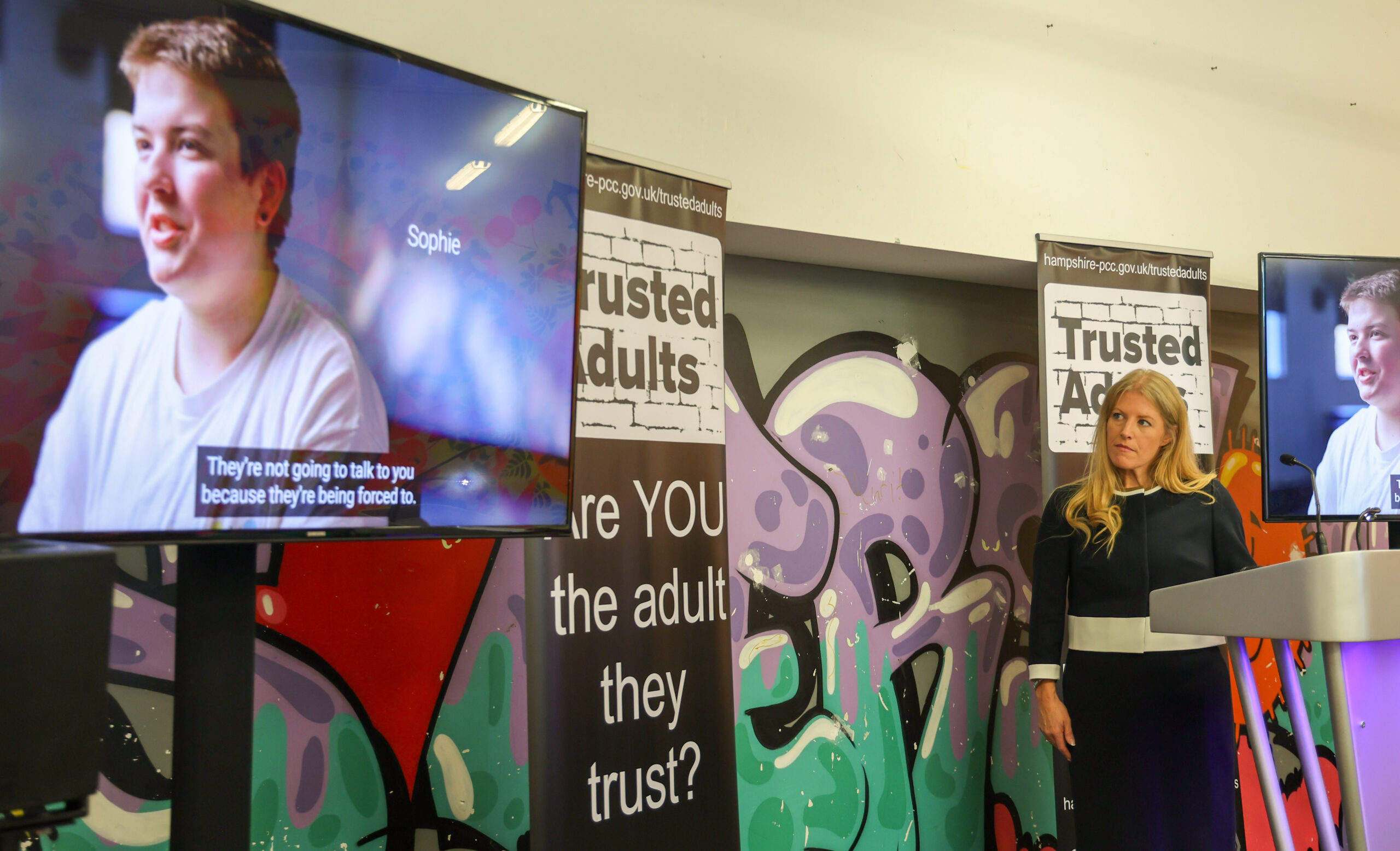 PCC Donna Jones watching a tv screen with a young person's image on it. The PCC is on a stage with two banners behind her, bearing the Trusted Adults branding.