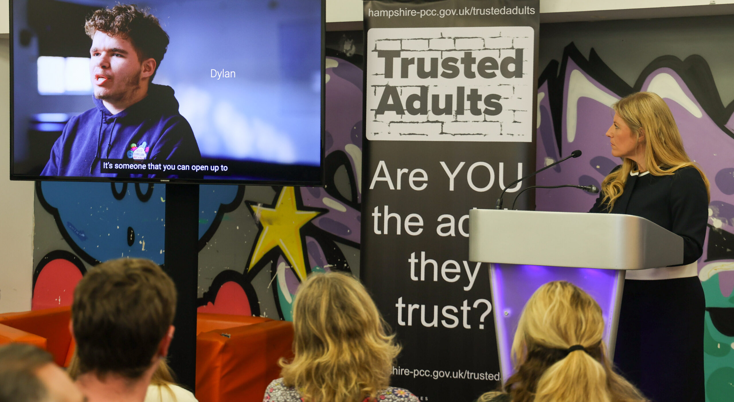 PCC Donna Jones watching a tv screen with a young person's image on it. The PCC is on a stage with two banners behind her, bearing the Trusted Adults branding.